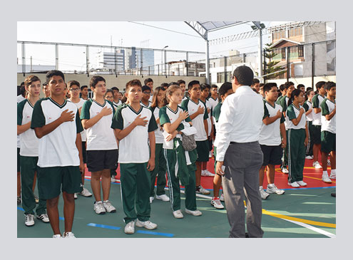 Alumnos con uniforme depostivo color verde con blanco con la mano en el pecho cantando himno